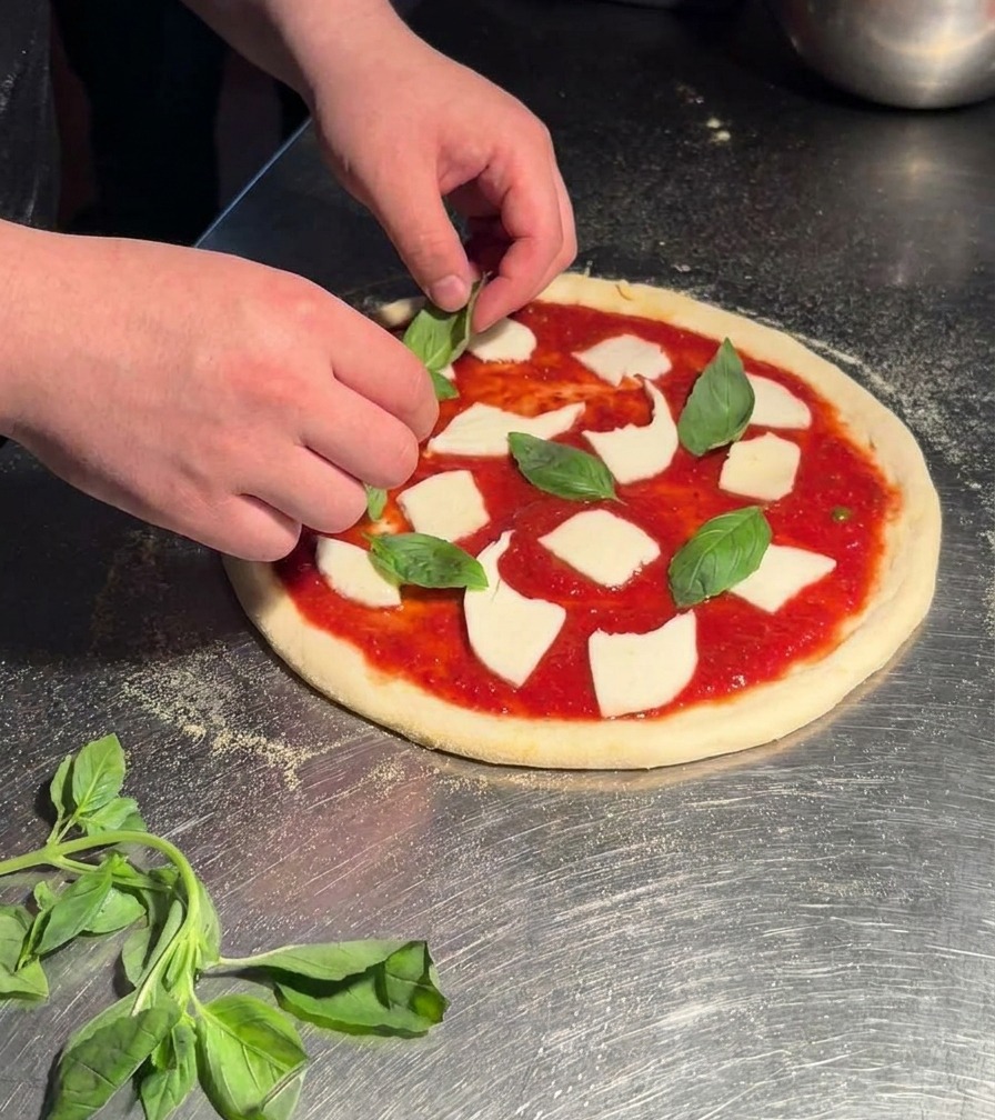 Des mains déposant des feuilles de basilic vert vif sur une pizza Margherita crue garnie de sauce et de mozzarella.