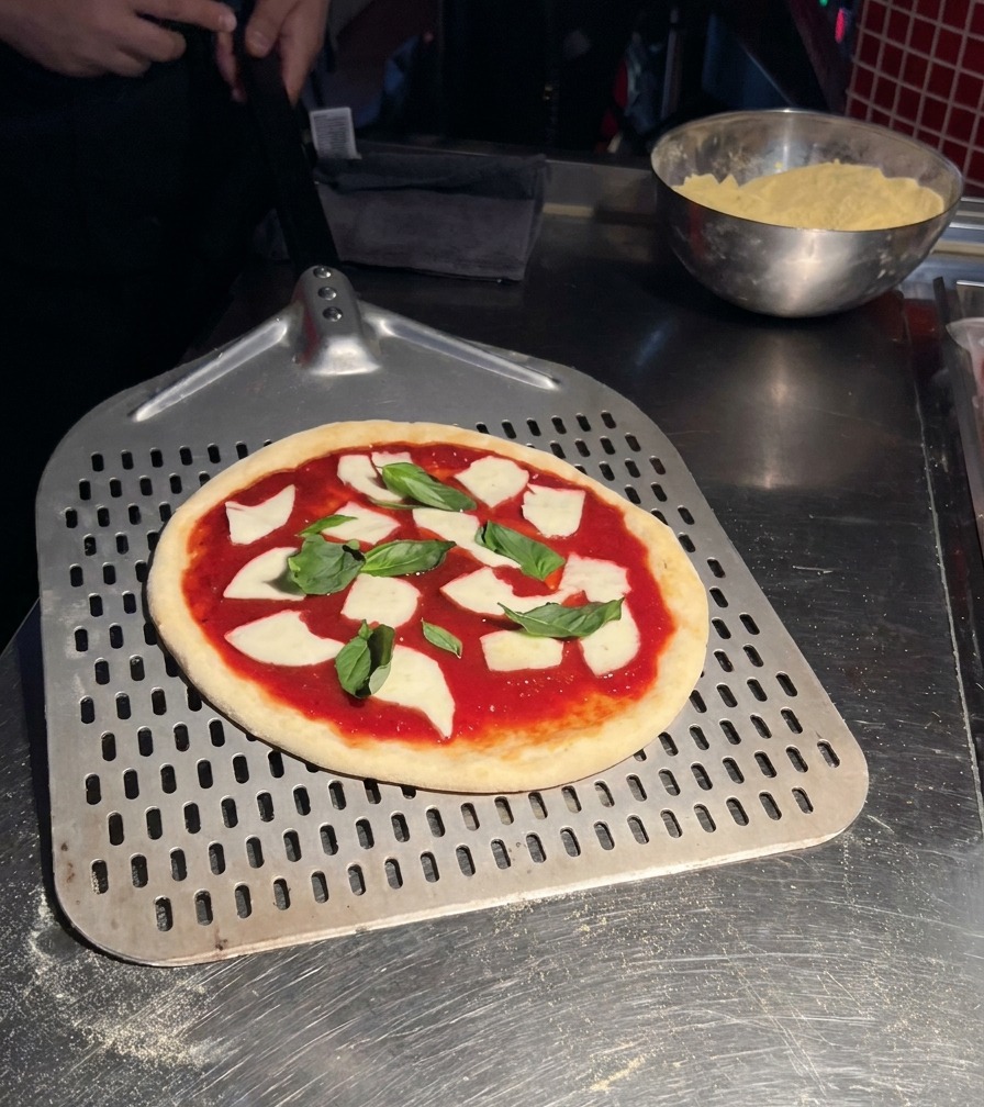 Une pizza Margherita crue garnie de sauce tomate, mozzarella et basilic posée sur une grande pelle à pizza en métal perforée.