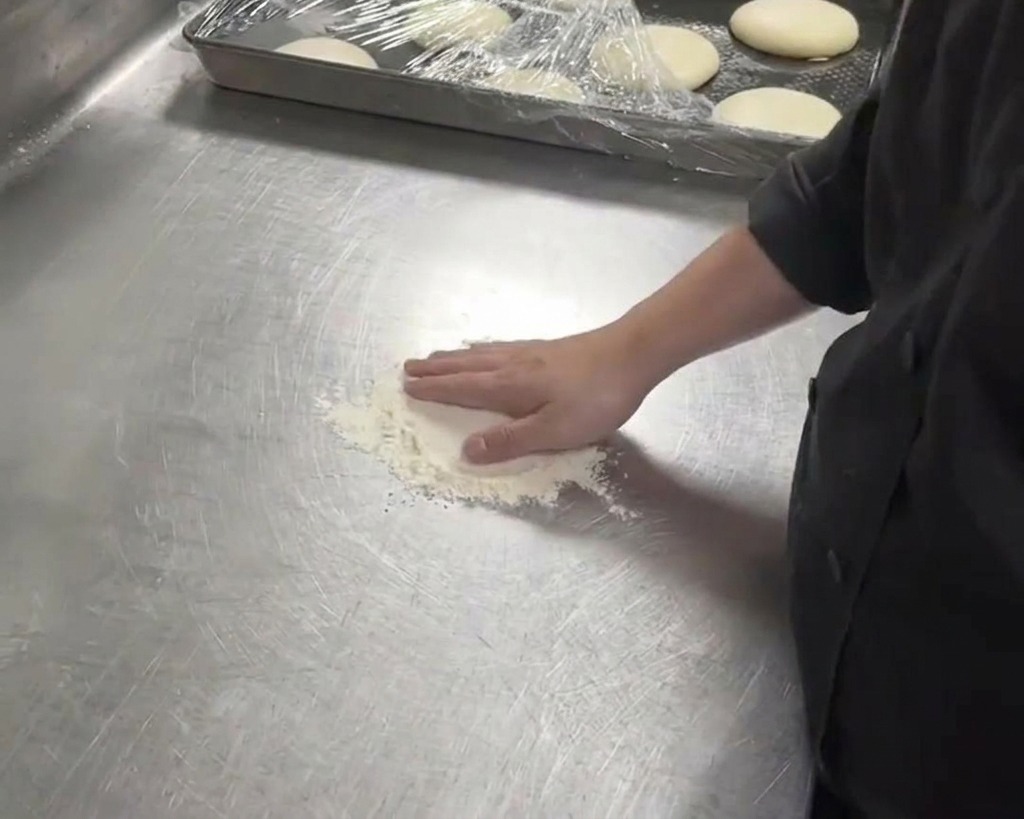 Une personne en veste de chef noire pressant sa main sur un pâton blanc sur un comptoir métallique fariné.