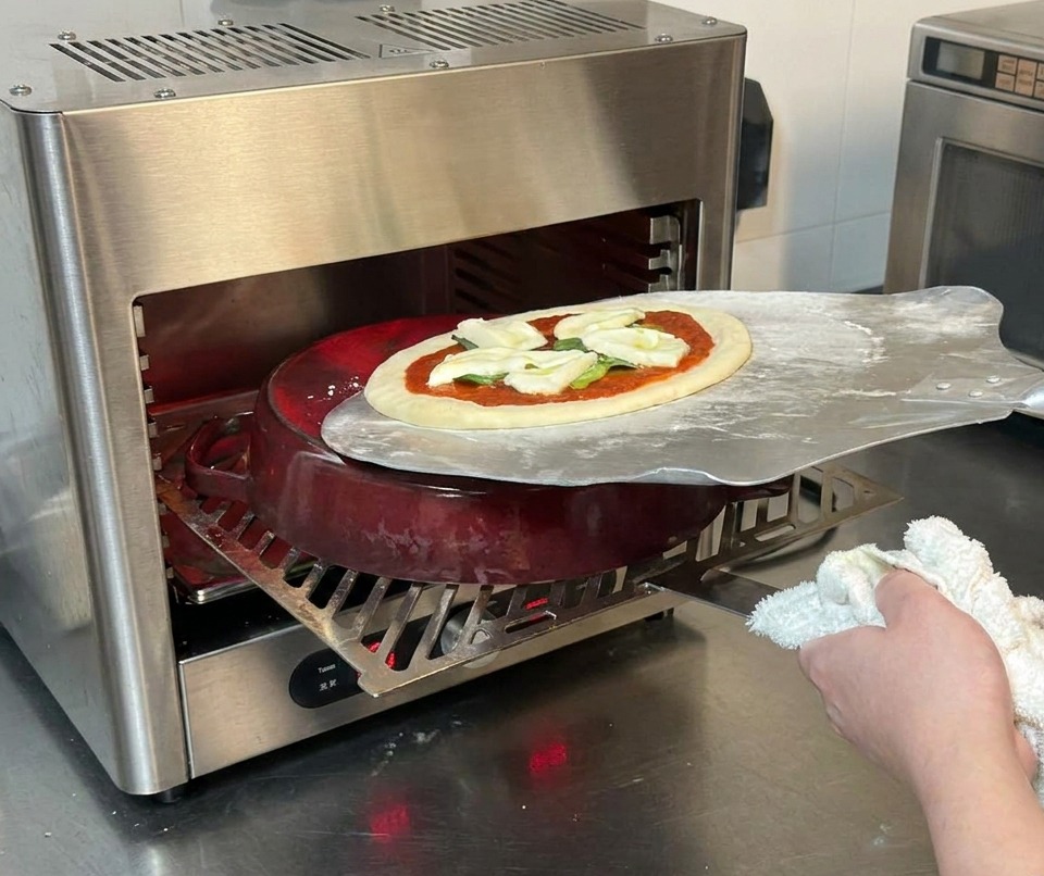Une pelle à pizza métallique faisant glisser une pizza crue sur une cocotte en fonte rouge à lintérieur dun four haute température.