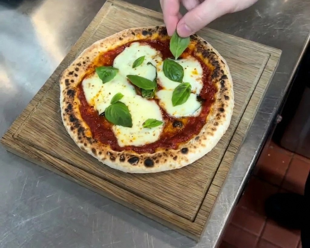 Une main posant délicatement des feuilles de basilic frais sur une pizza chaude sortant du four avec du fromage fondu et une croûte tachetée.