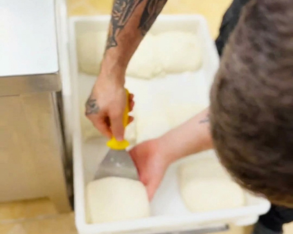 Une main de boulanger utilisant un coupe-pâte pour soulever délicatement un pâton blanc et souple dun bac en plastique.