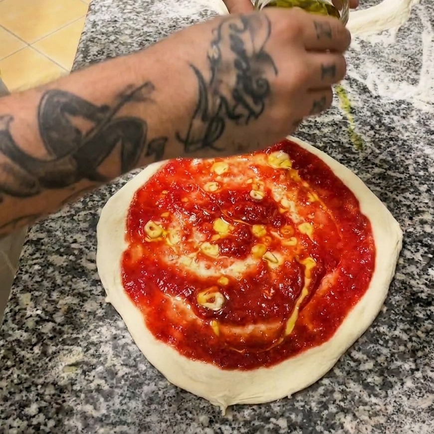 Une main utilisant une cuillère en métal pour étaler de la sauce tomate rouge vif en spirale sur la pâte crue.