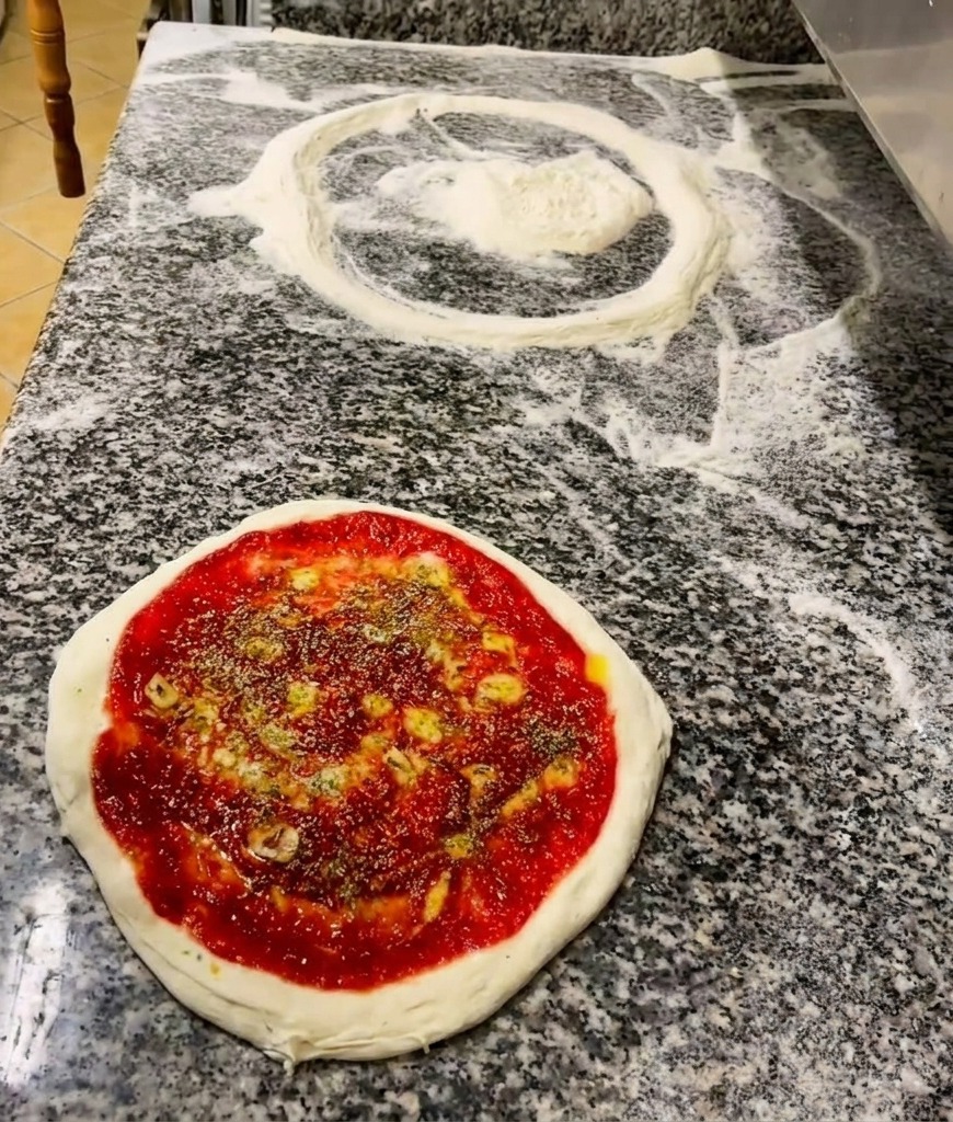 Une main saupoudrant des herbes séchées sur une base de pizza garnie de tomate et dun filet dhuile.