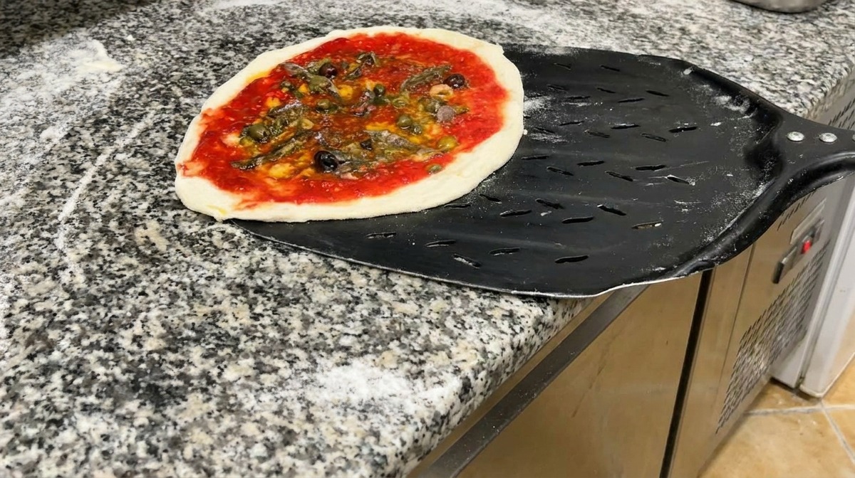 Une pizza prête à cuire posée sur une pelle à pizza en métal noir perforé.