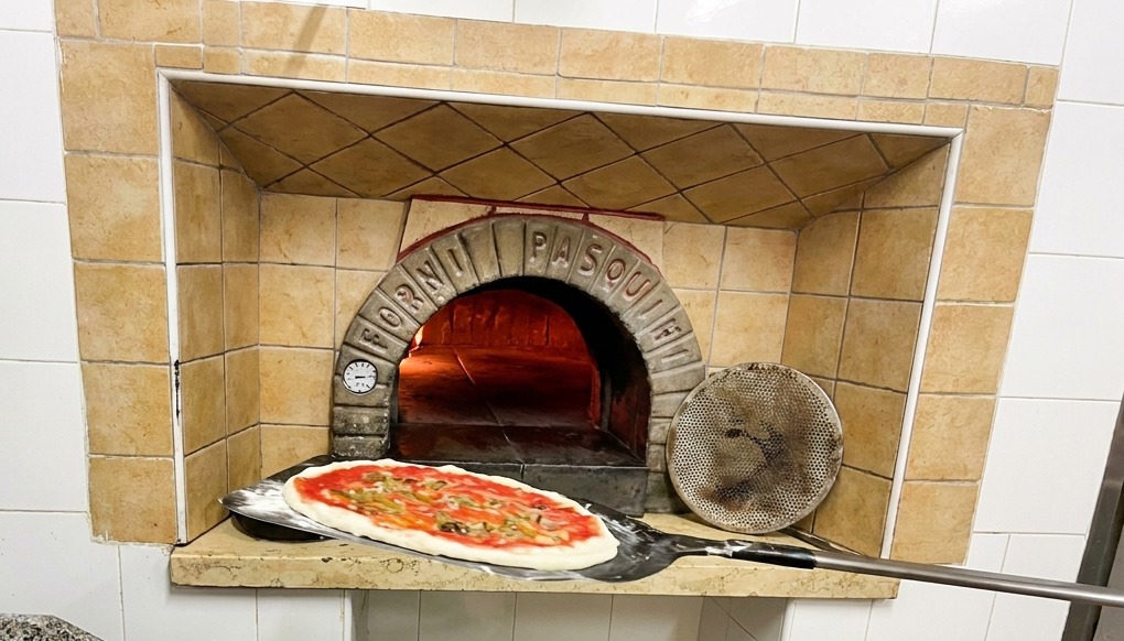 Une pizza sur une pelle à long manche insérée dans un four à bois traditionnel avec des flammes vives.