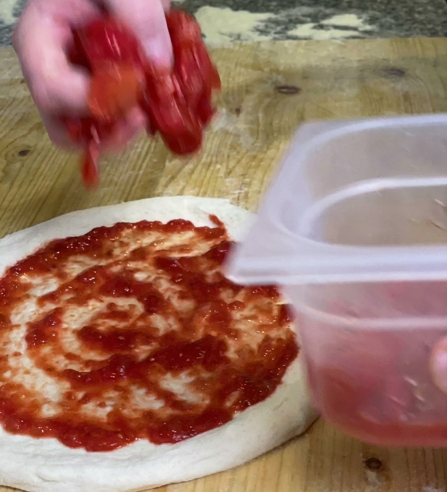 Une main déposant des morceaux de pulpe de tomate épaisse sur une base de pizza déjà nappée de sauce lisse.