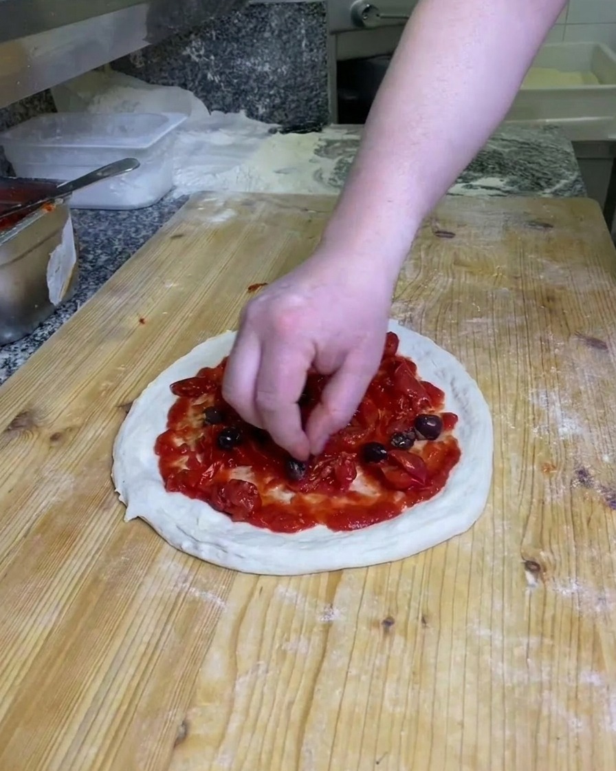 Une main plaçant des olives noires entières sur la pâte à pizza couverte de tomate sur un plan de travail fariné.