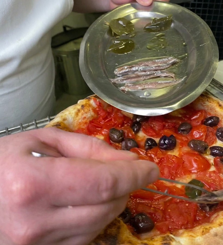 Un chef utilisant une pince pour déposer une feuille de laurier marinée sur une pizza garnie de tomates et dolives.
