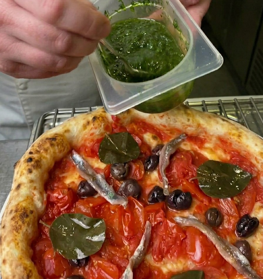 Un chef versant une sauce aux herbes vert vif depuis un contenant sur une pizza à laide dune petite cuillère.