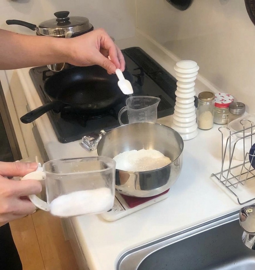 Une petite cuillère blanche ajoutant du sucre dans un bol de farine posé sur une balance de cuisine.