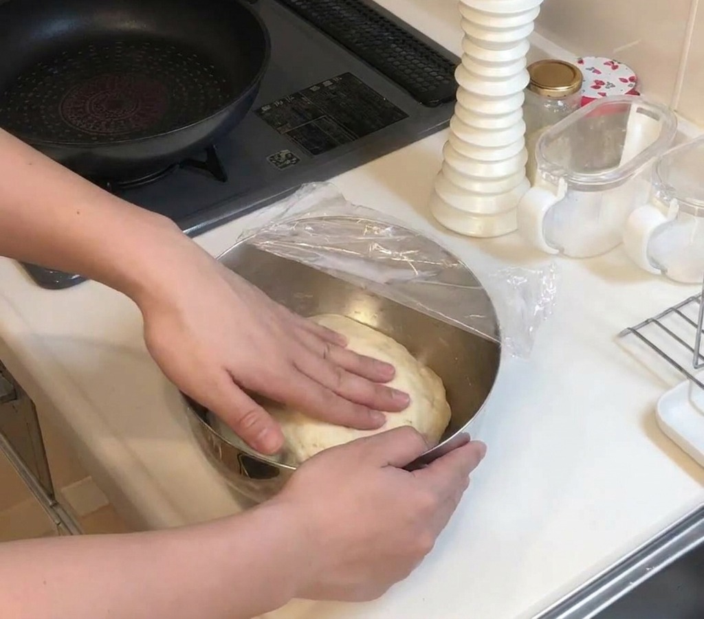 Des mains appuyant doucement sur une pâte levée dans un bol recouvert dun film plastique bien tendu.