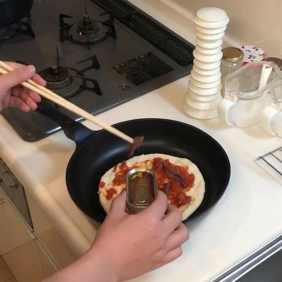 Des anchois sortis de leur boîte avec des baguettes et déposés sur le fond de pizza couvert de sauce.