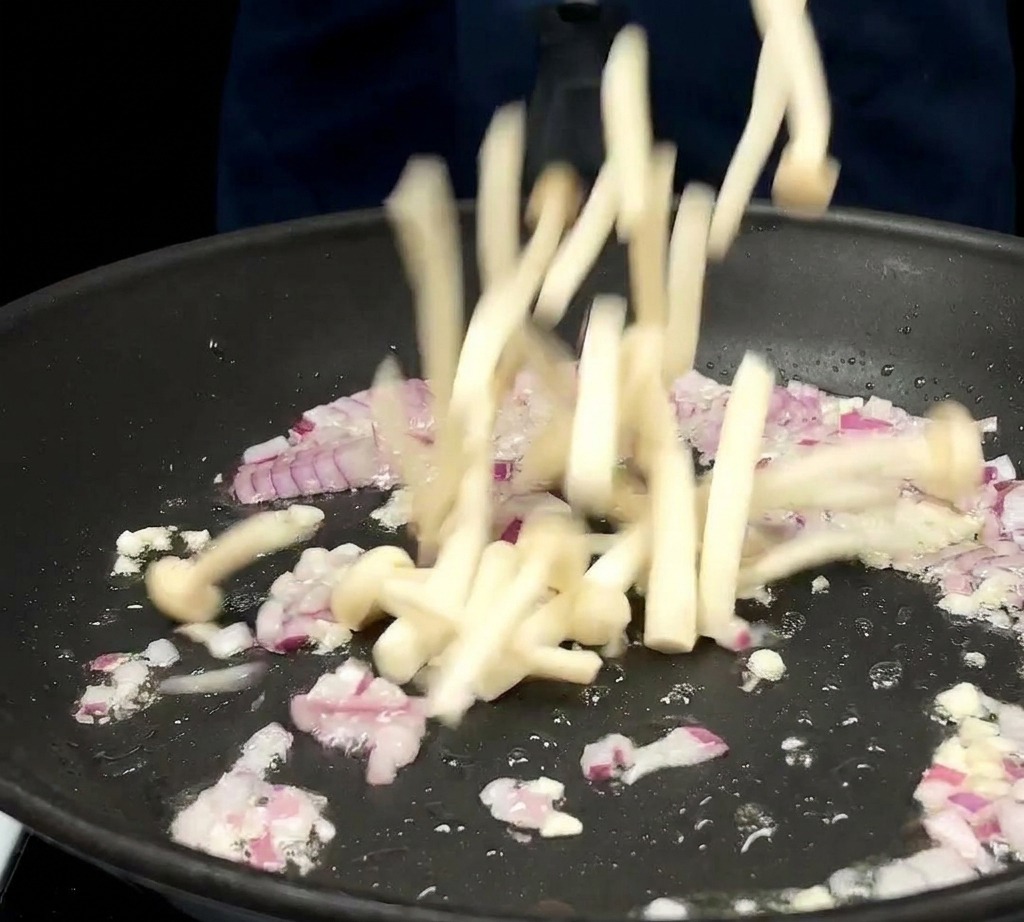 Une poignée de shimeji blancs versés dans une poêle contenant des oignons rouges et de lail sautés.