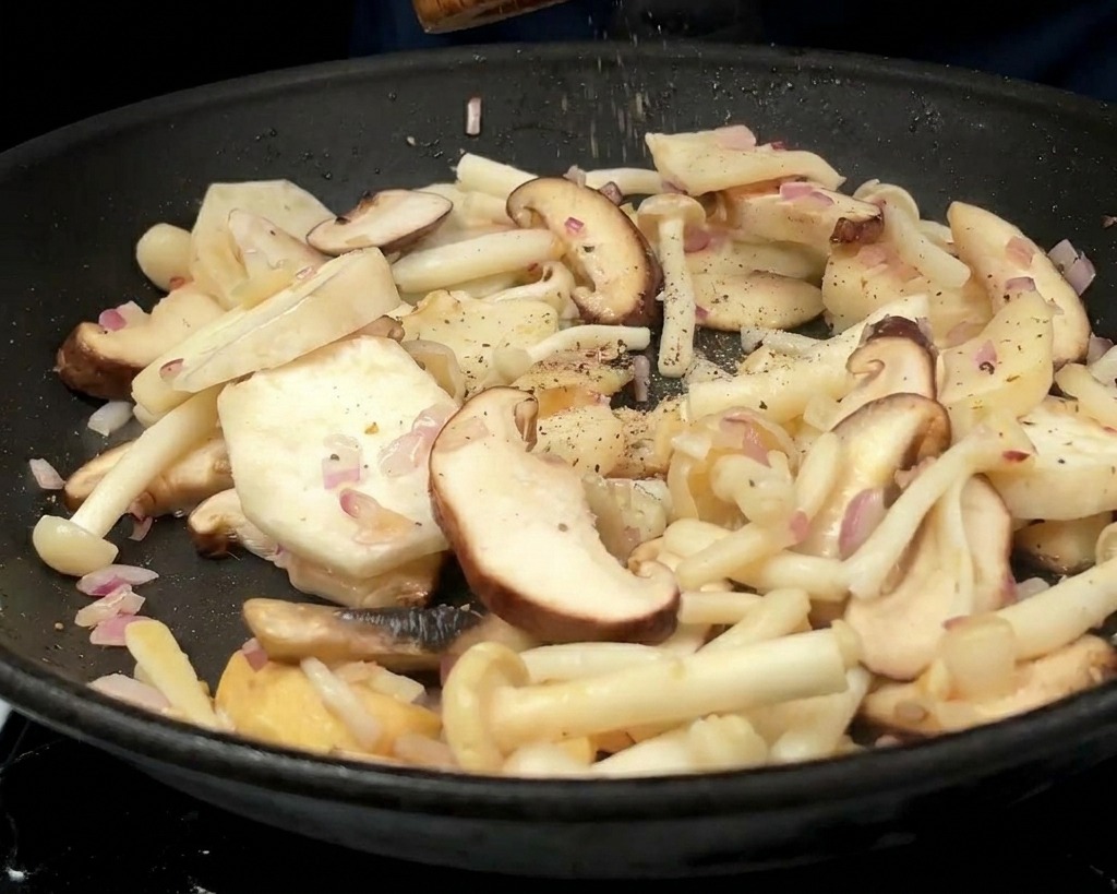 Une personne utilisant un moulin à poivre en bois pour moudre du poivre sur le mélange de champignons et doignons dans la poêle.
