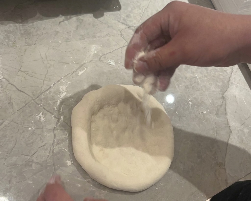 Mains pressant et aplatissant une boule de pâte crue sur un plan de travail en marbre gris saupoudré de farine blanche.