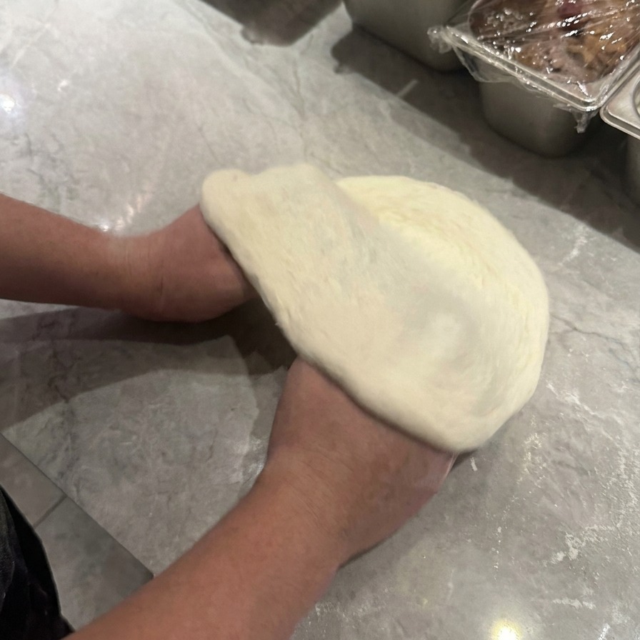 Deux mains soulevant et faisant pivoter la pâte à pizza crue pour létirer en cercle avec un bord extérieur visible.