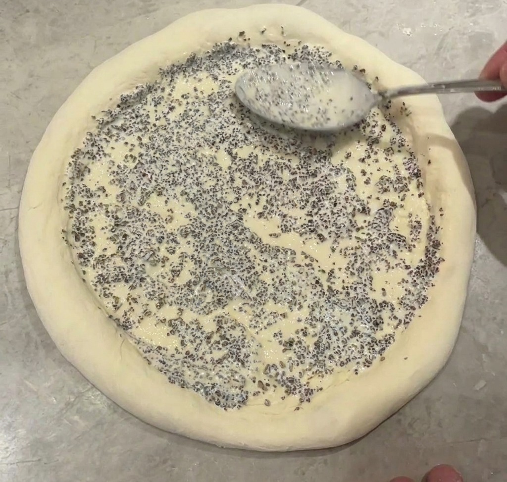 Une cuillère métallique étalant une sauce épaisse aux éclats de truffe noire sur le centre dune base de pizza crue.