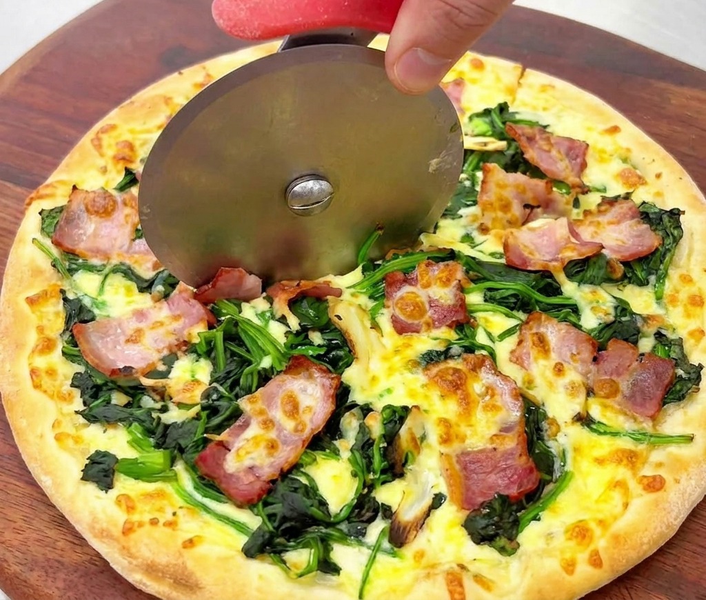 Gros plan dune roulette à pizza coupant une pizza dorée aux épinards et bacon croustillant.