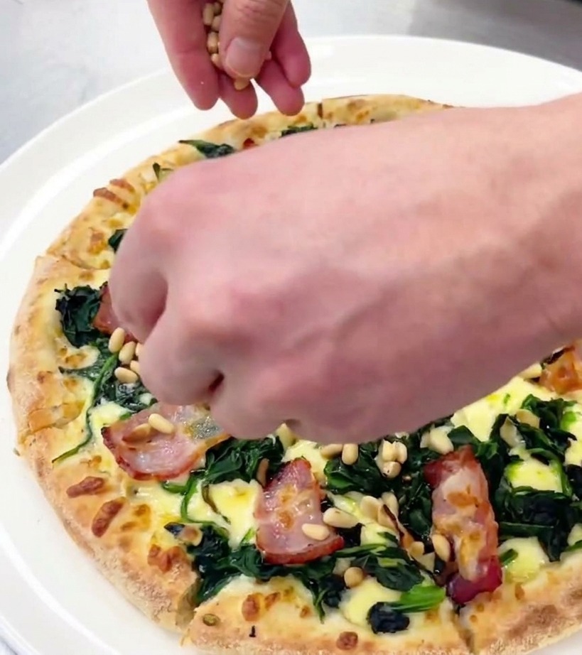Une main parsemant des pignons de pin grillés sur une pizza coupée présentée sur une assiette blanche.