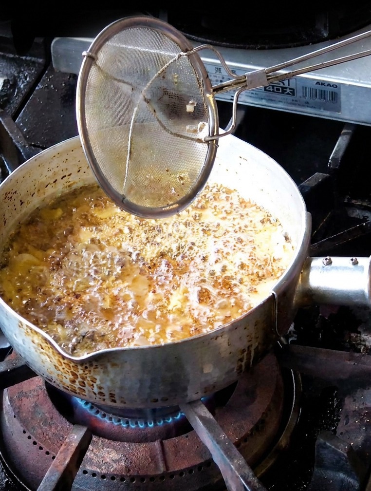 Une main utilisant une passoire pour retirer des tranches dail frit dun brun moyen dune casserole dhuile bouillonnante foncée.
