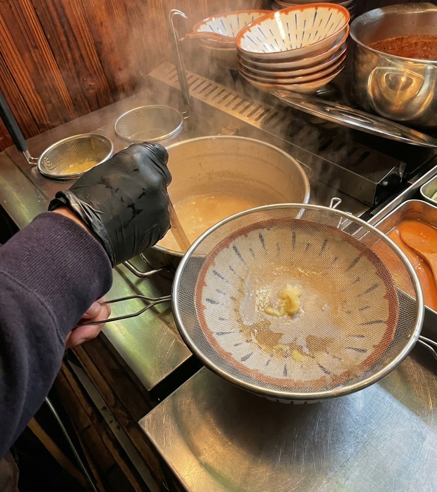 Bouillon dos crémeux versé à travers une passoire fine dans un bol à ramen japonais.