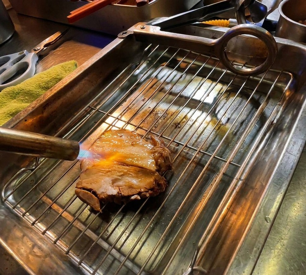 Un chalumeau de cuisine grillant une tranche épaisse de chashu de porc sur une grille métallique.