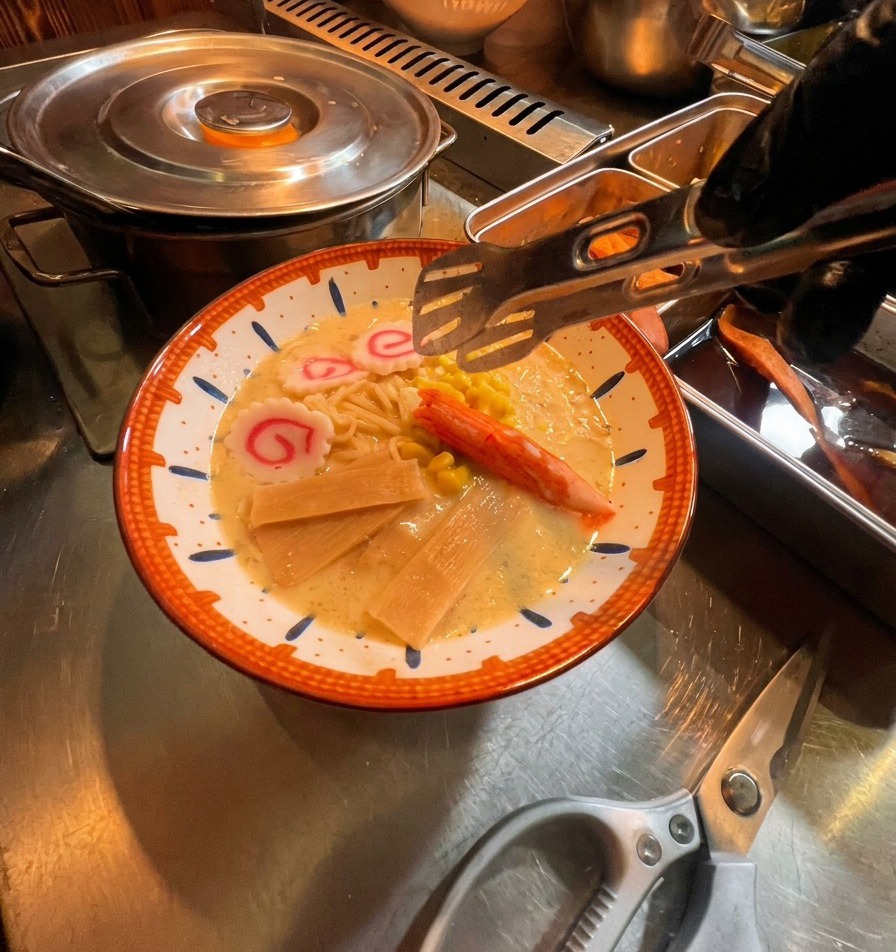 Une pince déposant du maïs, du surimi et des gâteaux de poisson sur le bol de ramen.
