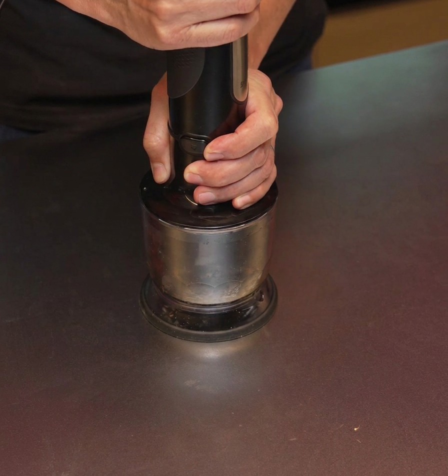 Une personne utilisant un mixeur plongeant noir pour broyer de lail carbonisé dans un récipient en métal.