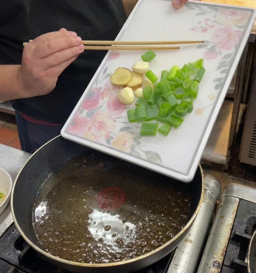 Des mains faisant glisser des oignons verts, du gingembre et de lail hachés dans une poêle dhuile chaude.