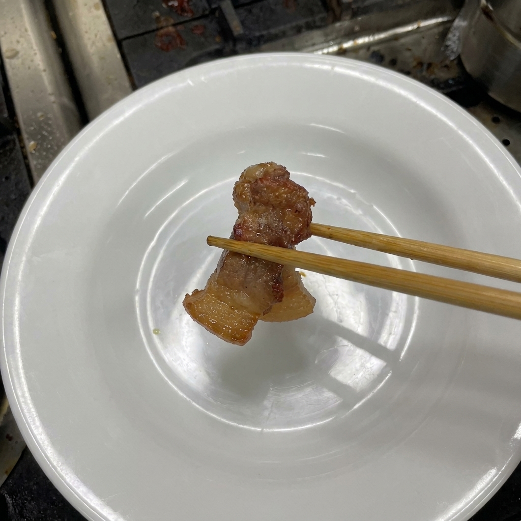 Une paire de baguettes en bois tenant un morceau de porc braisé tendre et brun foncé au-dessus dune assiette blanche.