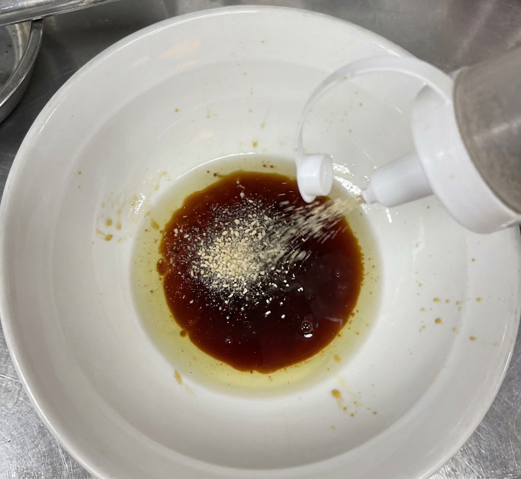 Poudre de sésame blanc saupoudrée dans un bol contenant une sauce brune et une huile claire.