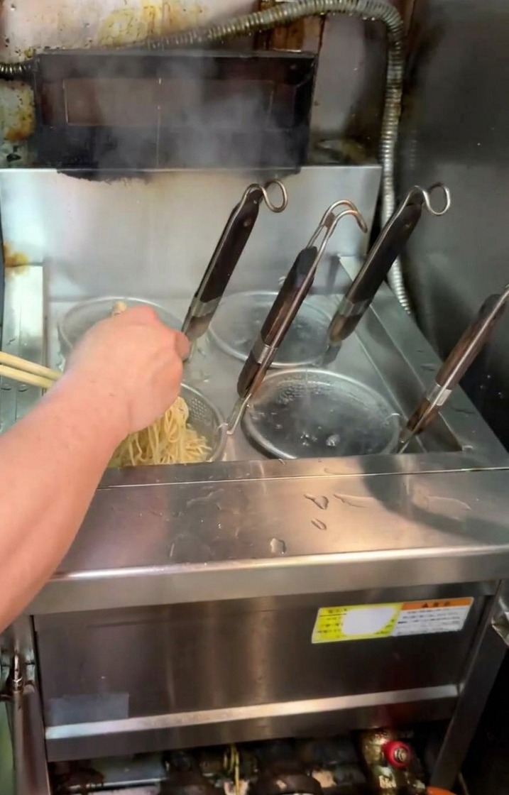 Un cuisinier soulevant une passoire à nouilles hors dune cuve de cuisson pour égoutter les nouilles ramen.