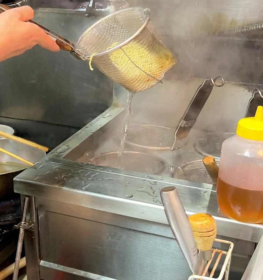 Une main tenant une passoire au-dessus dun bol, libérant les nouilles cuites dans un bouillon fumant.