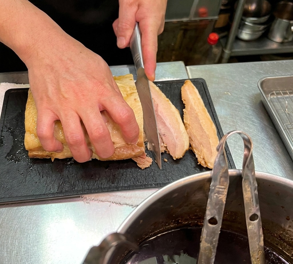 Gros plan dun chef coupant une pièce de poitrine de porc braisée en tranches sur une planche à découper noire.