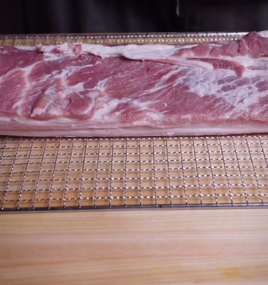 Un grand morceau rectangulaire de poitrine de porc crue sur une grille de refroidissement posée sur une planche à découper.