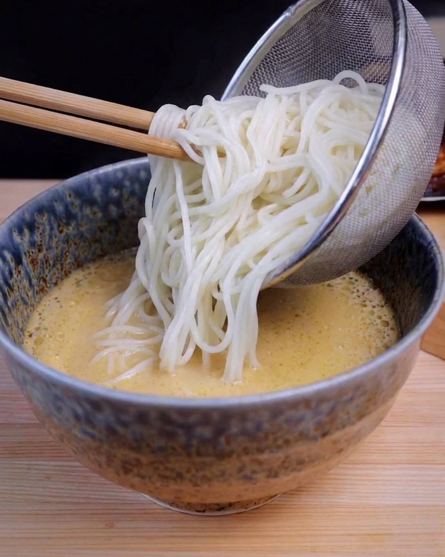 Une passoire en inox versant de longues nouilles blanches dans un bol contenant un bouillon ramen jaune.