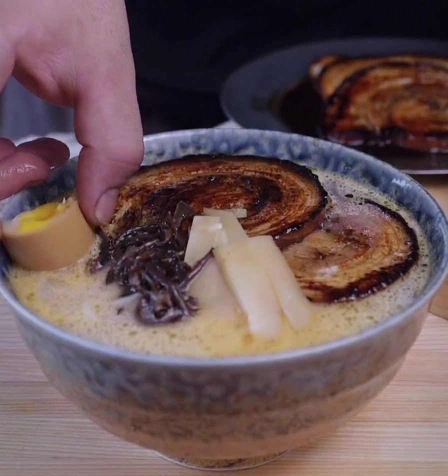 Une main déposant une moitié dœuf mollet dans un bol de ramen garni de chashu, de champignons et de bambou.