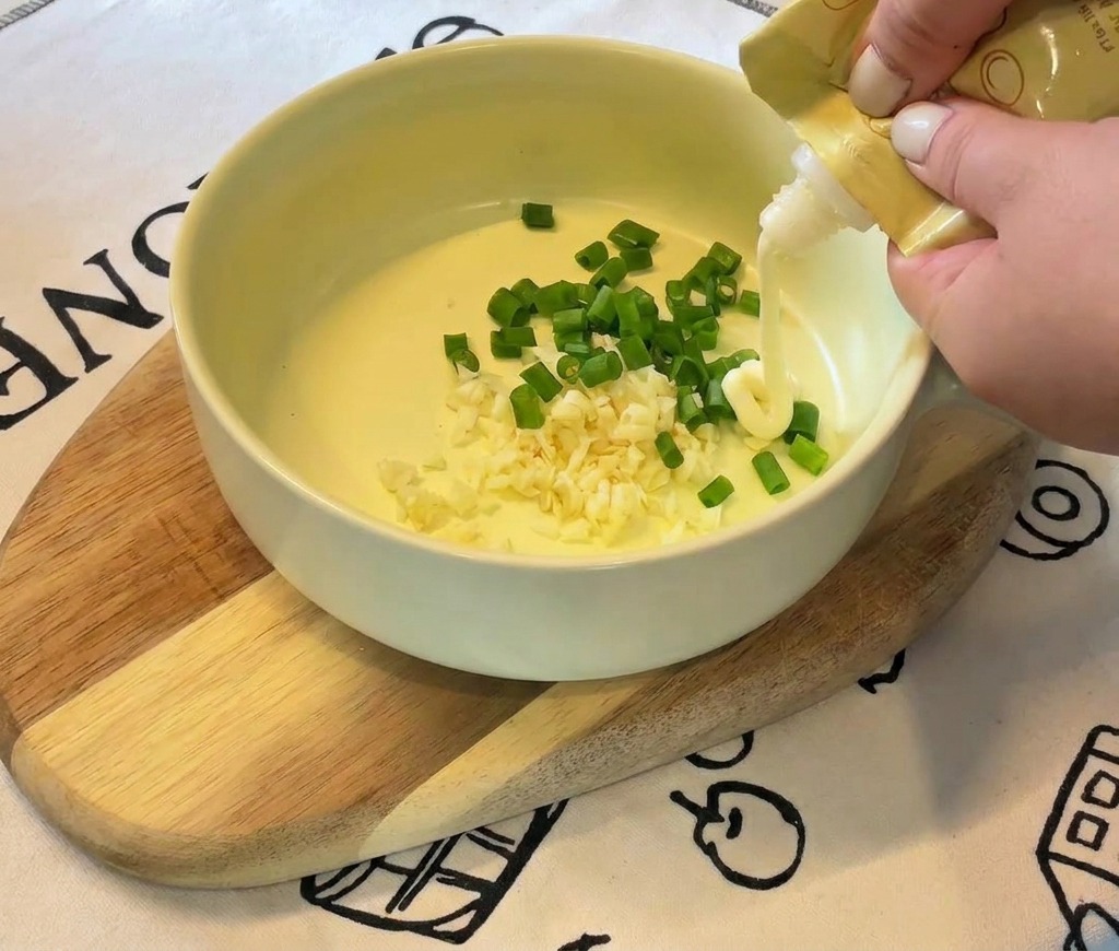 Les mains dune personne pressant de la mayonnaise blanche dun tube jaune dans un bol contenant de lail et des oignons verts.