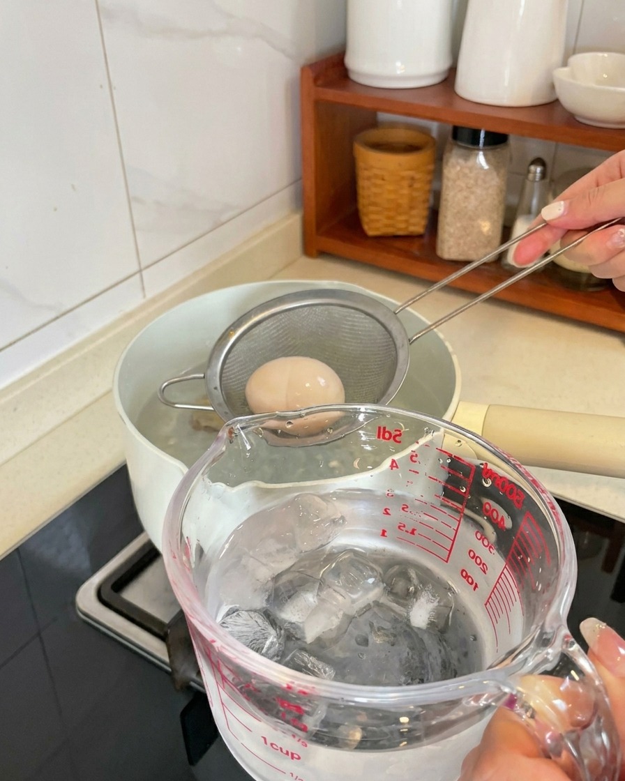 Un œuf dur soulevé dune casserole dans une passoire et plongé dans un verre doseur rempli de glaçons et deau.