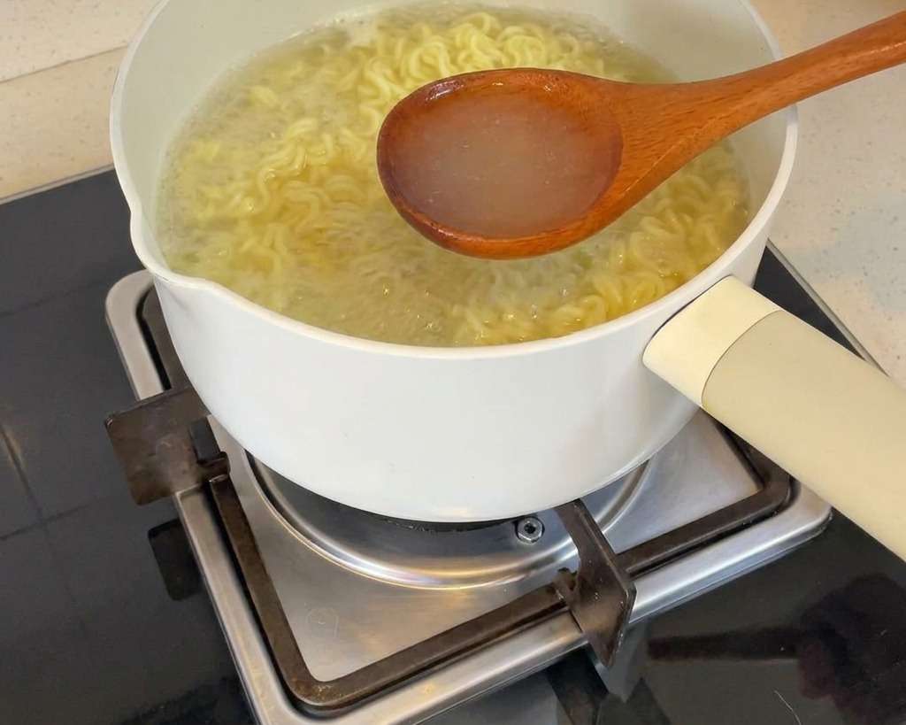 Une louche en bois prélevant leau de cuisson claire et fumante des nouilles dans une casserole sur le feu.