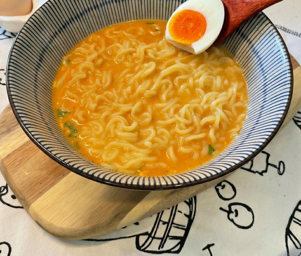 Une cuillère en bois déposant un demi-œuf mollet au jaune vif dans un bol de nouilles ramen au bouillon orangé.