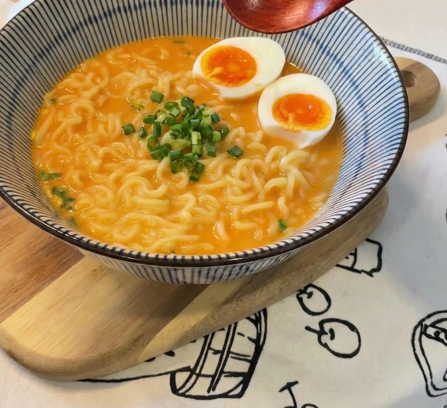 Un bol de ramen garni dœufs et dun petit tas doignons verts frais au centre, surmonté dune cuillère en bois.