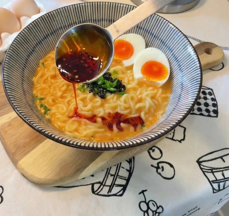 De lhuile pimentée rouge vif versée depuis une cuillère doseuse métallique sur un bol de ramen terminé avec des œufs, des oignons et des algues.