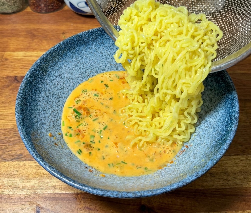 Des nouilles ramen jaunes cuites versées dune passoire en maille dans un bol bleu contenant un bouillon crémeux orangé.