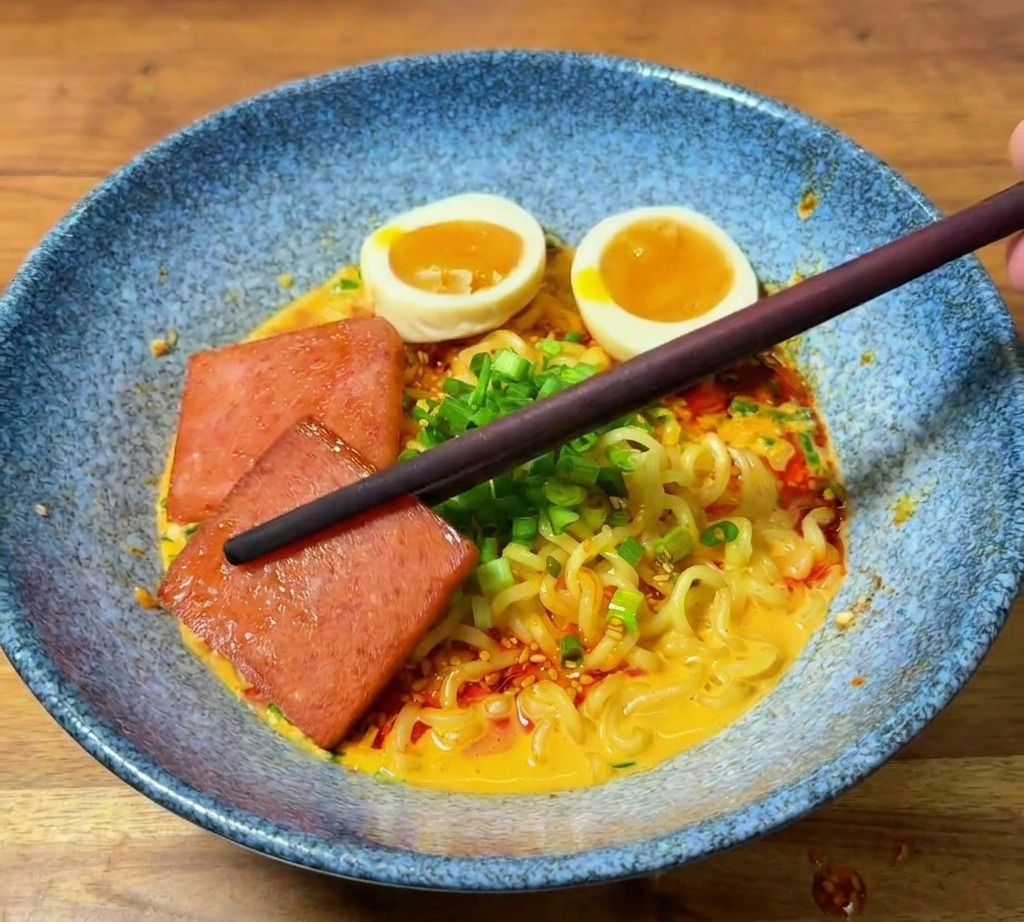 Des baguettes déposant un morceau de viande grillée dans un bol de ramen déjà garni dœufs et dhuile pimentée.