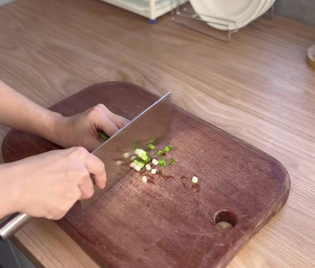 Une personne utilise un grand couteau de cuisine pour émincer finement des oignons verts sur une planche à découper en bois sombre.