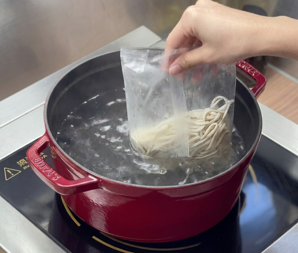 Des nouilles ramen fraîches sont versées dun sachet plastique transparent dans une casserole deau bouillante.