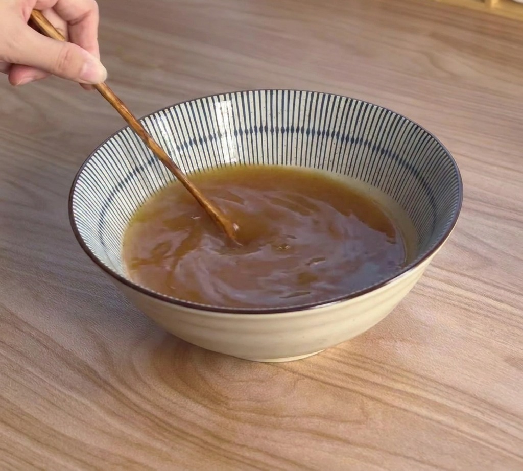 Une main utilise une cuillère en bois pour mélanger un bouillon miso brun dans un bol bleu et blanc.