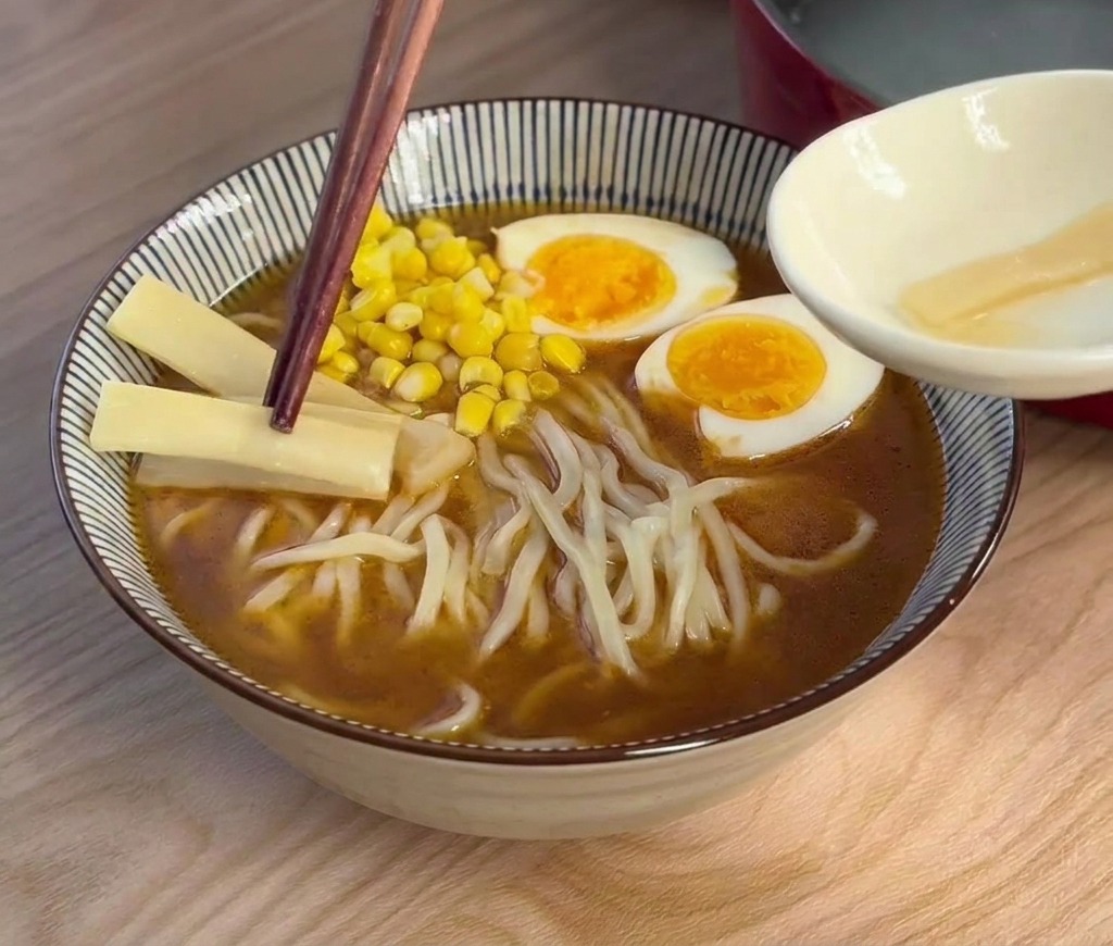 Des baguettes déposant de fines tranches de pousses de bambou claires dans un bol de ramen déjà garni.